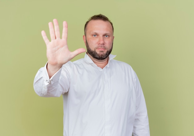 보여주는 흰 셔츠를 입고 수염 난된 남자가 가벼운 벽 위에 서있는 심각한 얼굴로보고 손가락 번호 5를 가리키는
