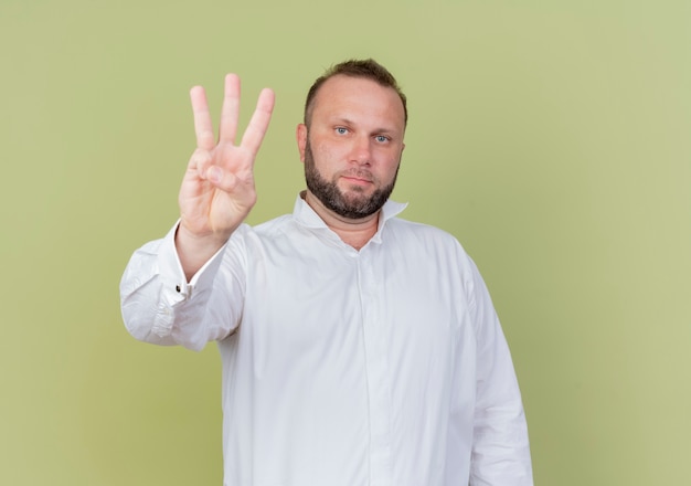 Бородатый мужчина в белой рубашке показывает пальцами номер три и смотрит вверх с серьезным лицом, стоящим над светлой стеной