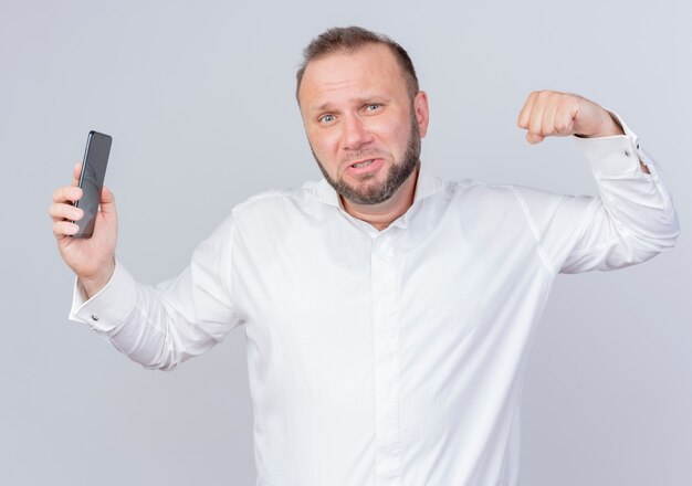 Bearded man wearing white shirt showing smartphone clenching fist showing biceps looking confident standing over white wall