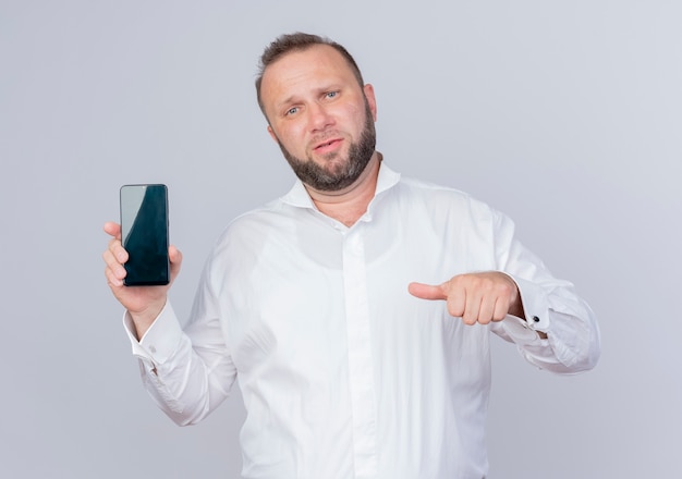 Bearded man wearing white shirt showing smartphone pointing with thumb at it looking displeased standing over white wall