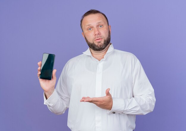 Bearded man wearing white shirt showing smartphone presenting with arm standing over blue wall