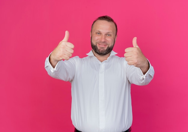 Uomo barbuto che indossa una camicia bianca che mostra i pollici in su sorridendo allegramente in piedi sopra il muro rosa