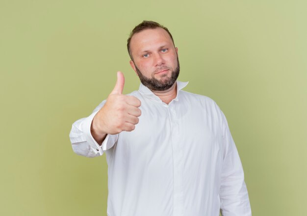 Бородатый мужчина в белой рубашке показывает палец вверх, уверенно улыбаясь, стоя над светлой стеной