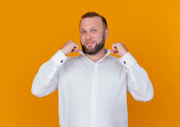 Bearded man wearing white shirt  smiling confident fixing his collar standing over orange wall