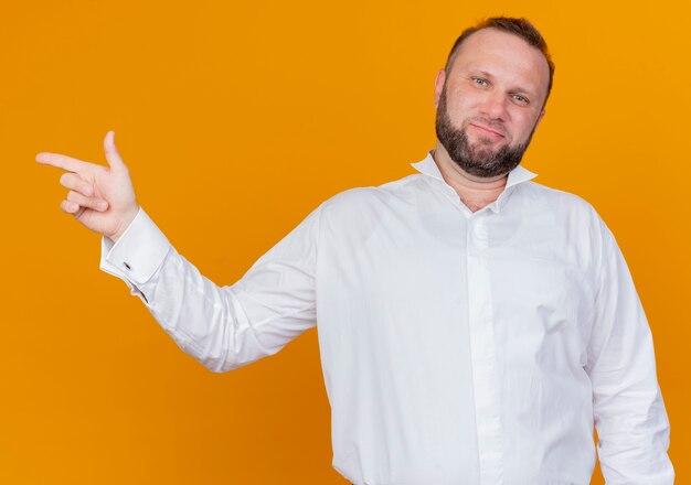 Bearded man wearing white shirt  smiling pointing with index finger to the side standing over orange wall