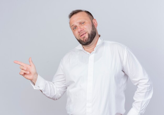 Bearded man wearing white shirt  smiling pointing with index finger to the side standing over white wall