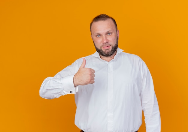 Bearded man wearing white shirt  smiling showing thumbs up standing over orange wall