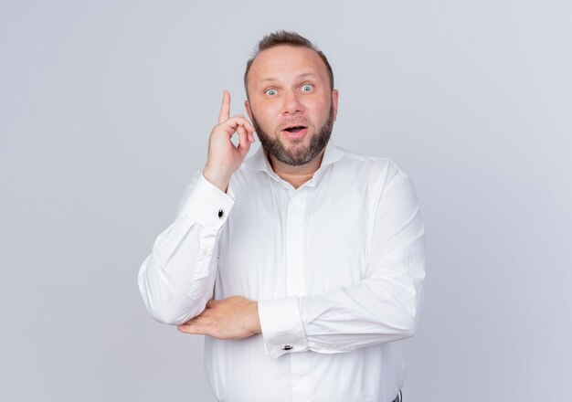 Bearded man wearing white shirt  smiling surprised showing index finger having great new idea standing over white wall
