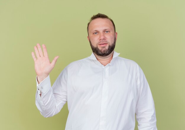 Bearded man wearing white shirt  smiling waving with hand standing over light wall