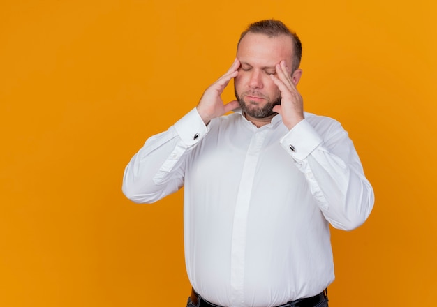 Foto gratuita uomo barbuto che indossa una camicia bianca che tocca la sua testa che soffre di mal di testa in piedi sopra la parete arancione