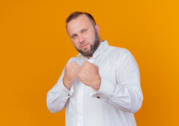 Bearded man wearing white shirt  with clenched fists posing like boxer standing over orange wall