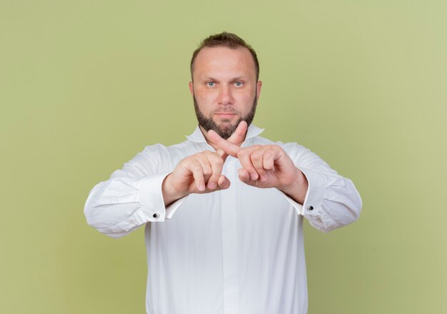 Free photo bearded man wearing white shirt  with serious face crossing index fingers standing over light wall