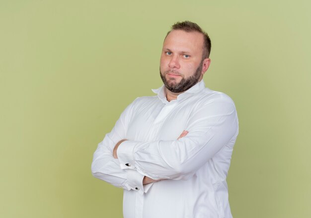 Bearded man wearing white shirt  with serious face with arms crossed on chest standing over light wall