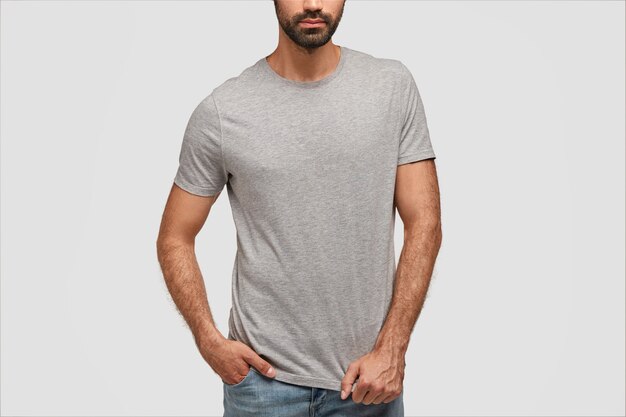 ひげを生やした男は空白のグレットTシャツとジーンズを着て、コンクリートの壁に立っています