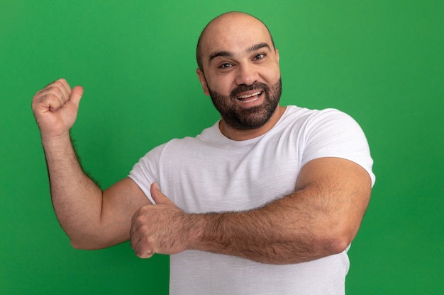Bearded man in white t-shirt  happy and cheerful pointing with fingers back standing over green wall