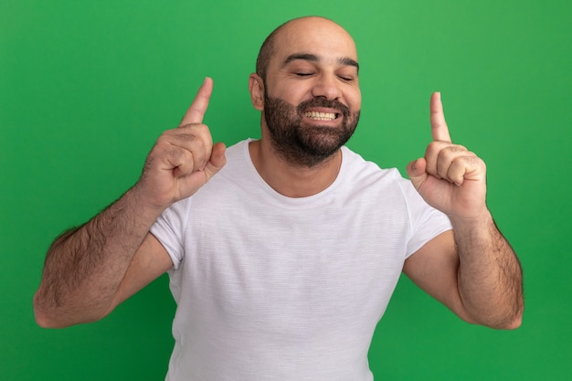 흰색 티셔츠 행복하고 쾌활한 수염 난 남자가 녹색 벽 위에 서있는 검지 손가락을 광범위하게 보여주는 미소