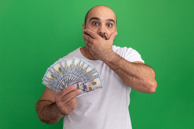 Бородатый мужчина в белой футболке, держащий деньги в шоке, прикрывает рот рукой, стоящей над зеленой стеной