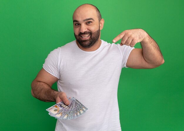 Bearded man in white t-shirt holding cash happy and positive pointing with index finger at money standing over green wall