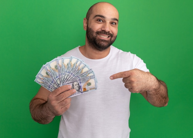 Bearded man in white t-shirt holding cash  happy and positive smiling cheerfully pointing with index finger at money standing over green wall