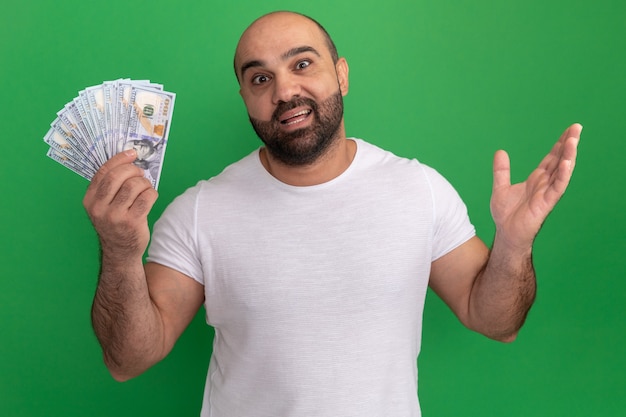 Бородатый мужчина в белой футболке держит деньги счастливым и удивленным с поднятой рукой, стоя над зеленой стеной