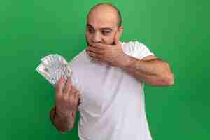 Free photo bearded man in white t-shirt holding cash looking at money amazed and surprised covering mouth with hand standing over green wall