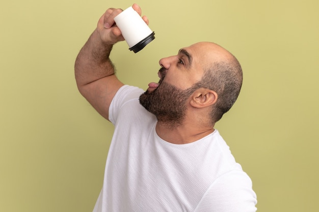 Бородатый мужчина в белой футболке держит чашку кофе, собираясь пить, стоя у зеленой стены