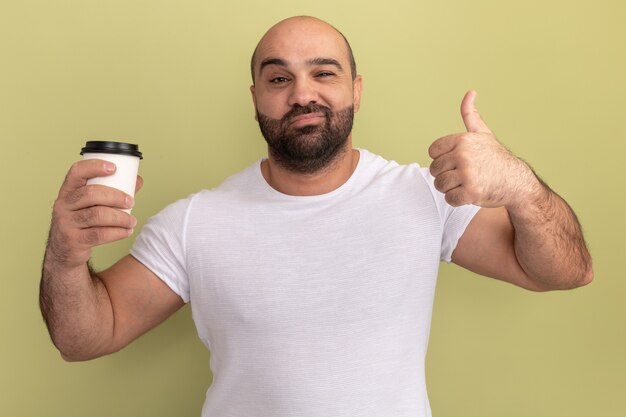 緑の壁の上に幸せで前向きなコーヒーカップを保持している白いTシャツのひげを生やした男