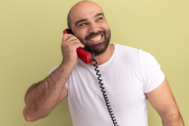 Bearded man in white t-shirt holding old telephone looking aside with big smile on face standing over green wall