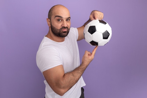 Bearded man in white t-shirt holding soccer ball on his finger  with serious face standing over purple wall