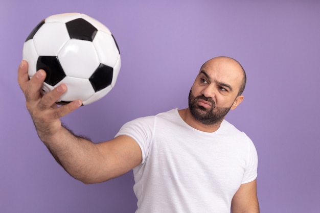 Bearded man in white t-shirt holding soccer ball looking at it with skeptic expression standing over purple wall