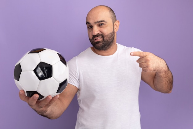 Bearded man in white t-shirt holding soccer ball  with confident expression pointing with index finger to the side standing over purple wall