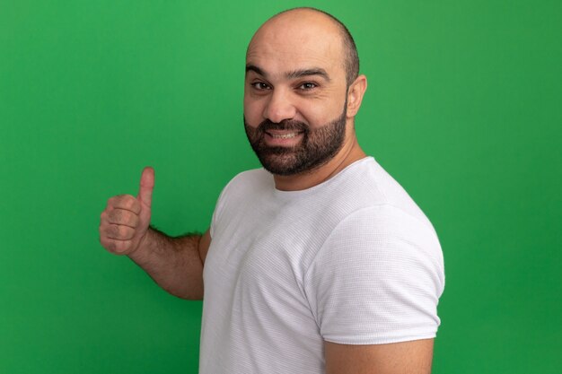 Bearded man in white t-shirt  smiling confident showing thumbs up standing over green wall