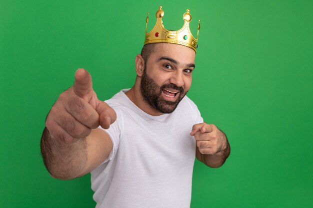 Bearded man in white t-shirt wearing golden crown  happy and positive pointing with index fingers standing over green wall