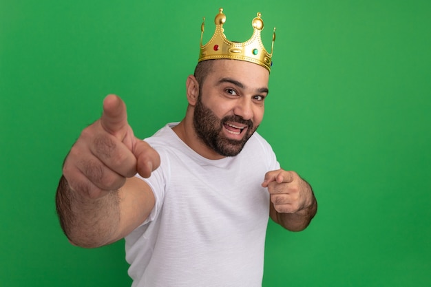 Бородатый мужчина в белой футболке с золотой короной счастлив и позитивно показывает указательными пальцами, стоя над зеленой стеной
