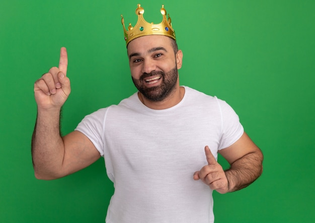 Бородатый мужчина в белой футболке с золотой короной счастлив и позитивно показывает указательными пальцами вверх, стоя над зеленой стеной