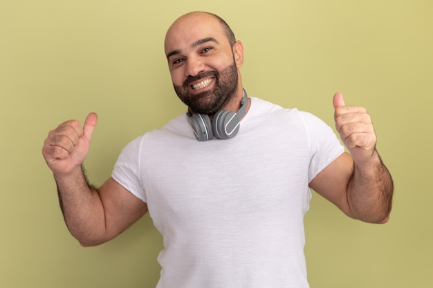 Bearded man in white t-shirt with headphones  happy and cheerful smiling showing thumbs up standing over green wall