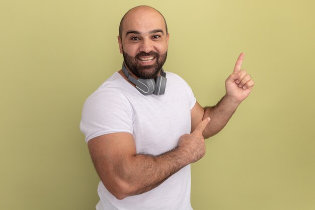 Бородатый мужчина в белой футболке с наушниками счастливый и позитивный, указывая указательными пальцами в сторону, стоя над зеленой стеной