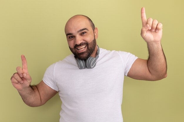 Бородатый мужчина в белой футболке с наушниками счастлив и позитивно улыбается, весело указывая указательными пальцами вверх, стоя над зеленой стеной