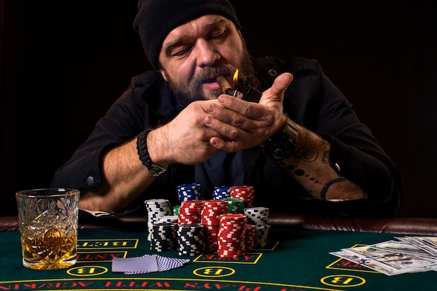 Free photo bearded man with cigar and glass sitting at poker table in a casino. gambling, playing cards and roulette. on the green poker table are cards, chips and money. the whole room is in smoke from cigars