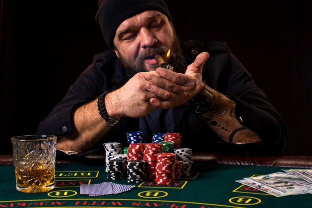 bearded man with cigar and glass sitting at poker table in a casino. gambling, playing cards and roulette. on the green poker table are cards, chips and money. the whole room is in smoke from cigars