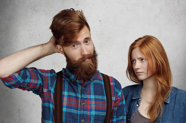 Bearded man with confused and guilty look, scratching his head while his redhead girlfriend standing next to him and looking with contempt and disappointement.