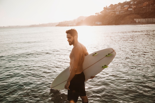 Uomo barbuto con tavola da surf sulla riva vicino all'acqua in tempo soleggiato