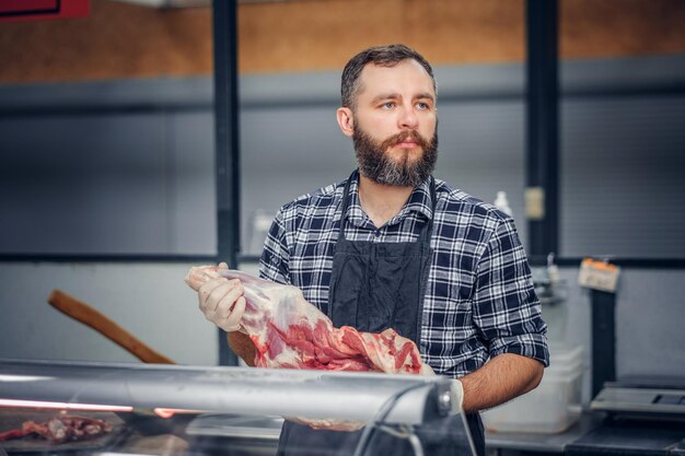 Бородатый продавец мяса, одетый в флисовую рубашку, продает на рынке свежее мясо.