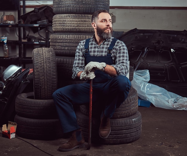 Foto gratuita meccanico barbuto vestito con un'uniforme, tiene un piede di porco, si siede su vecchi pneumatici per auto nel garage.