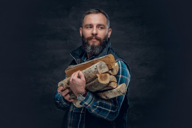 A bearded middle age male dressed in plaid flannel shirt holds firewoods over a dark background.