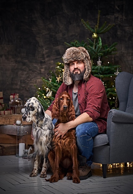 Бородатый мужчина средних лет, одетый в клетчатую рубашку и меховую шапку, и две ирландские сеттер-собаки с елкой и рождественским украшением на заднем плане.