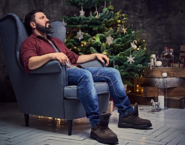 A bearded middle age male dressed in a plaid shirt and jeans sits on a chair over Christmas illumination and fir tree in background.
