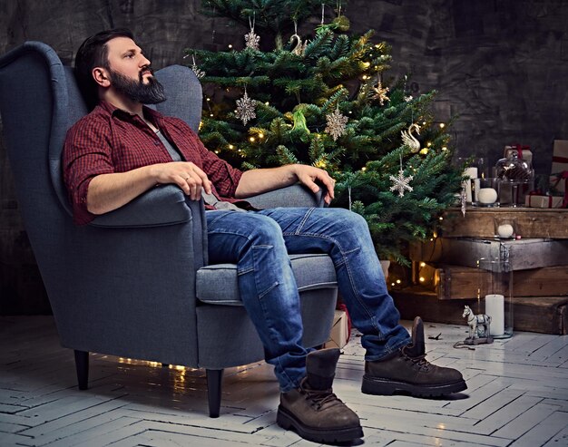Бородатый мужчина средних лет, одетый в клетчатую рубашку и джинсы, сидит на стуле над рождественской иллюминацией и елкой на заднем плане.