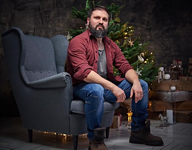 A bearded middle age male dressed in a plaid shirt and jeans sits on a chair over Christmas illumination and fir tree in background.