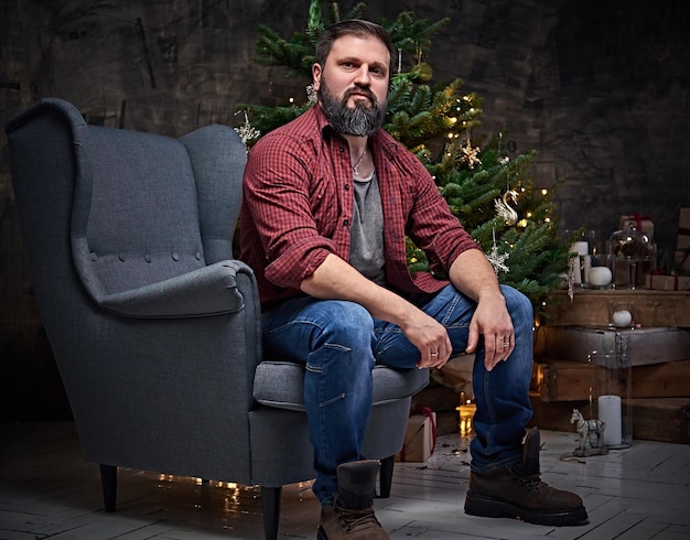 格子縞のシャツとジーンズに身を包んだひげを生やした中年男性は、クリスマスのイルミネーションと背景のモミの木の上に椅子に座っています。